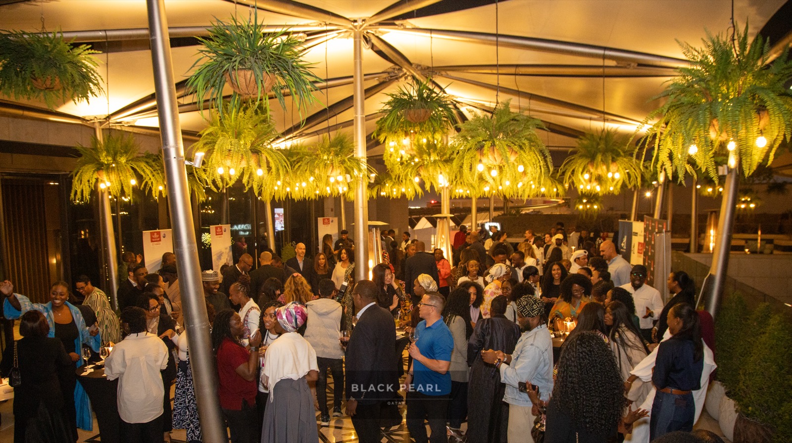A wide rooftop crowd of Black Pearl guests in warm conversation under string lights, Doha skyline beyond