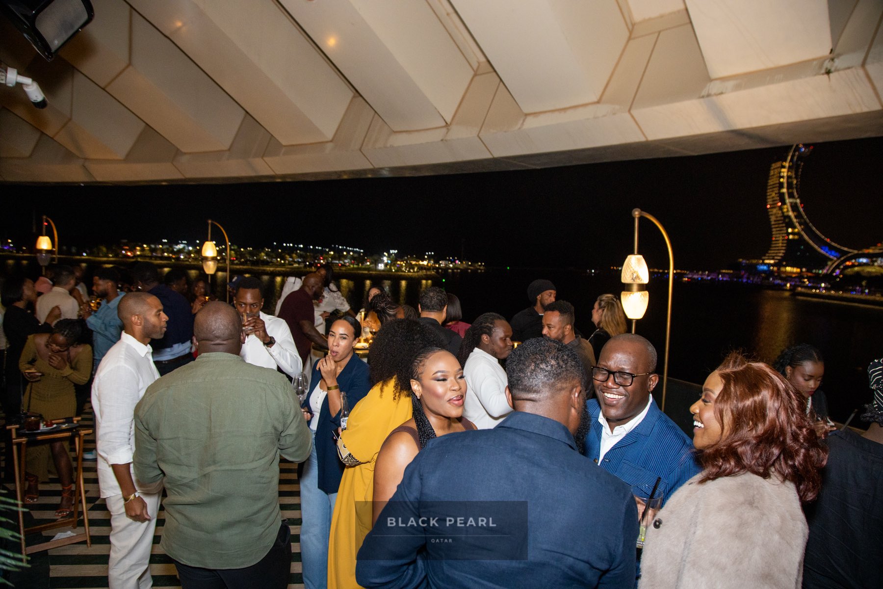 A crowded rooftop mixer at night with Doha's skyline lit up beyond the terrace